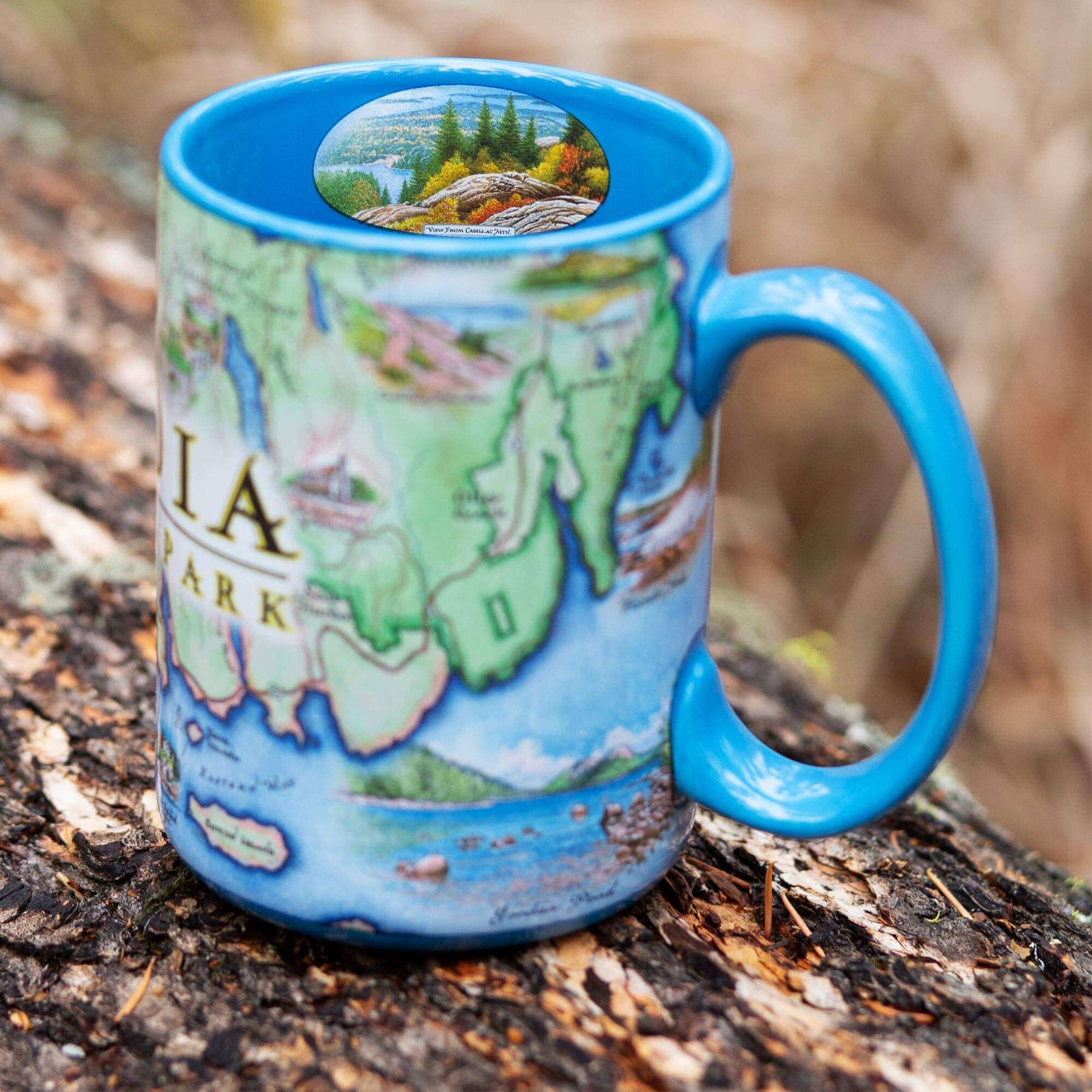 Maine's Acadia National Park Map Ceramic Mug in the color blue sitting on a log.  The coffee cup features Cadillac Mountains, Otter Cliffs, Beaver, and Jordon Pond. 