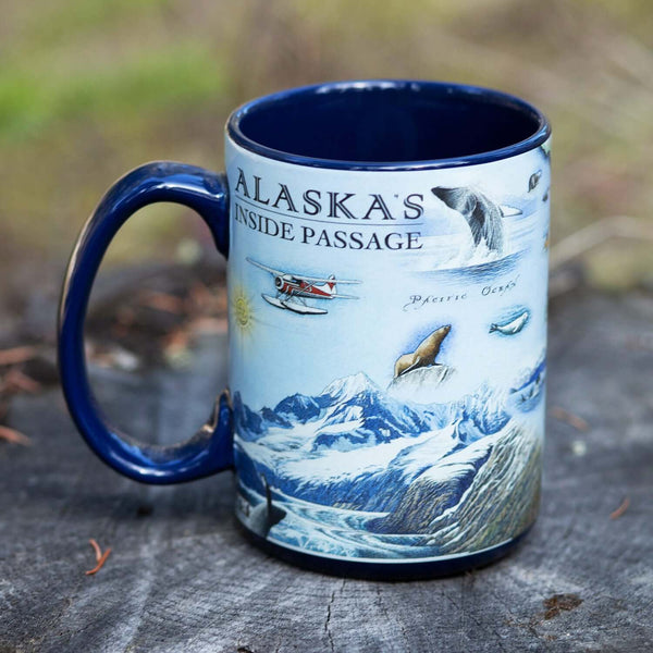 Blue Alaska's Inside Passage Mug sitting on a log with the coffee cup featuring a pontoon airplane, whale jumping out of water, and mountains.