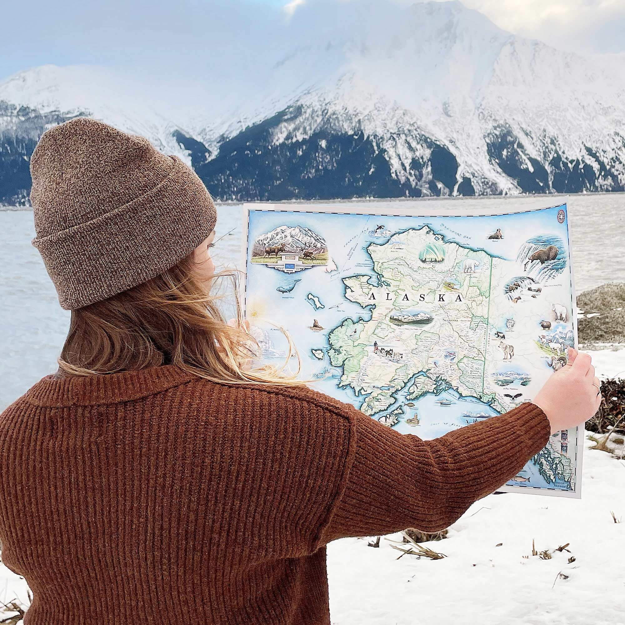 A woman is holding up the Alaska State Xplorer Map in front of mountains on the Turnagain Arm of the Seward Highway.