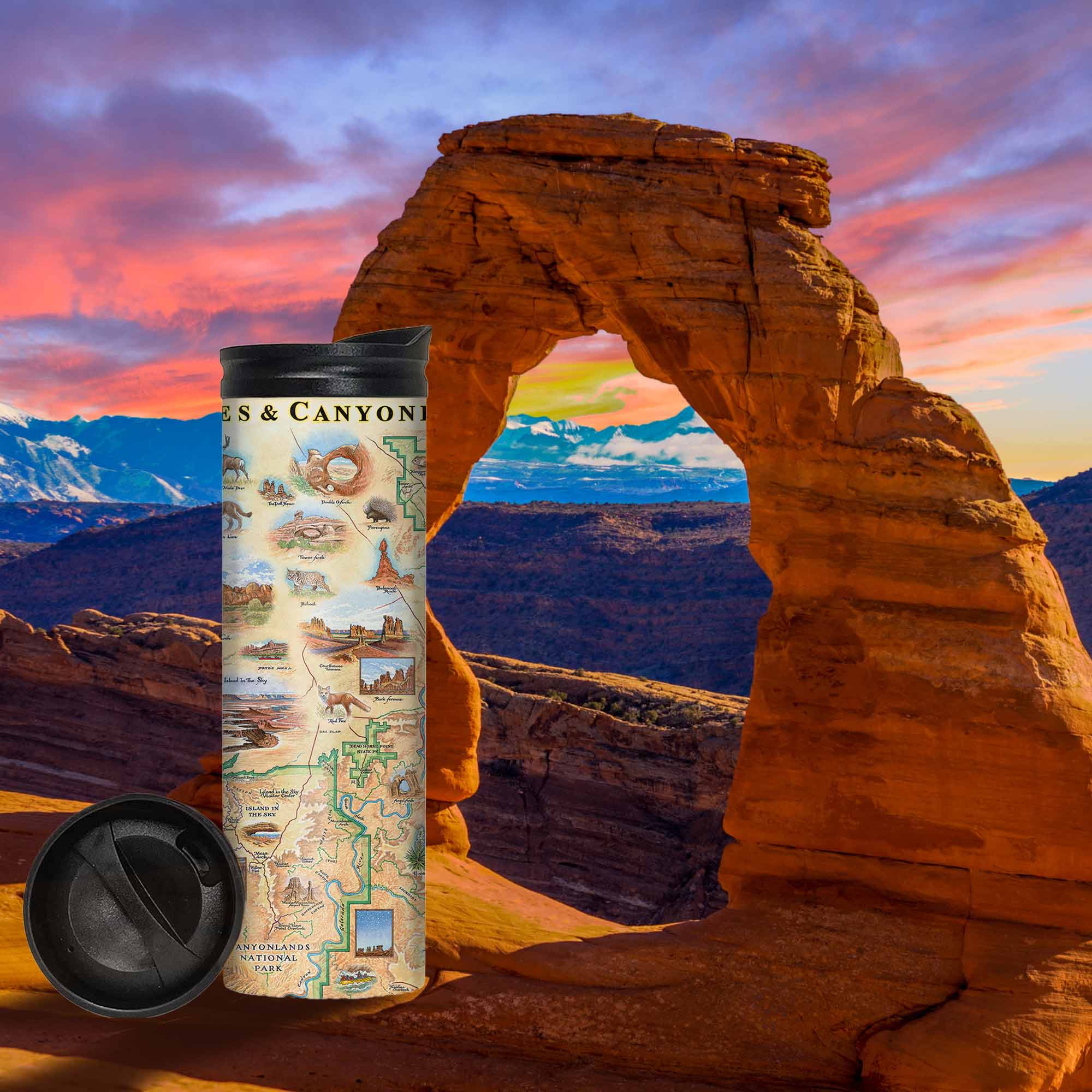 Utah's Arches & Canyonlands National Parks Map Travel Drinkware - 16 oz sitting in front of Delicate Arch during a sunset or sunrise. Utah's Arches & Canyonlands National Parks map travel Drinkware in earth tone colors of blues and greens. The map features deer, mountain lion, porcupine, bobcats, and a fox. Landmarks are Tower Arch Delicate Arch, Double O Arch, Balanced Rock, Island in the Sky, and Fiery Furnace. 