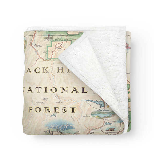 Black Hills National Forest Map Fleece Blanket in earth-tone colors. The map features Mount Rushmore, Jewel Cave National Monument, Custer State Park, and Devil's Tower. The map also includes flora and fauna of areas such as Bison, Elk, Big Horn Sheep, Bald Eagle, Rocky Mountain Iris, and Buffalo Berries. 