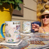 Boston, Massachusetts city map ceramic mug sitting on a table with coaster. A women is in the background drinking coffee. The cup features Boston Strong, the Boston Marathon - Blue - 16 oz.