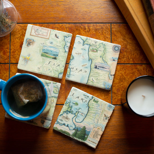 Montana's Flathead Lake Natural Stone Coaster Set of 4 by Xplorer Maps. The coasters are sitting on a tile counter top with a mug full of tea. Features Golfing, Steamboat, Rafting, birds, eagles, Osprey, Bears, forest, water, deer, mountain lion, fish, Cherry Blossoms, and flowers like Glacier Lilies and Lady Slippers. Cities and landmarks are noted such as Woods Bay, Wild Horse Island, Finley Point, and Big Arm. 