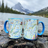 Montana's Glacier National Park Ceramic Coffee Mug sitting on a rock in the river with mountains in the background. Cups featuring mountains, Goats, flowers, and more! Blue - 16 oz.
