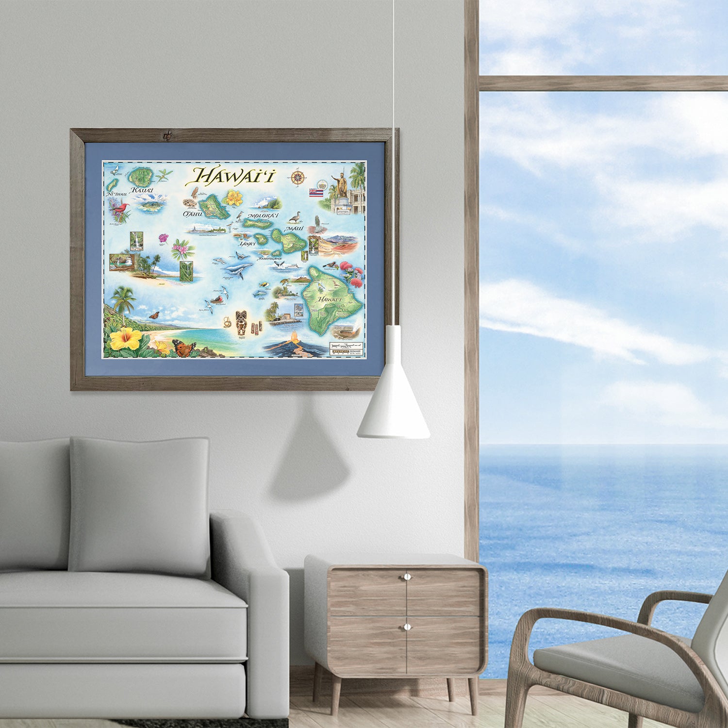 Xplorer Maps state map of Hawaii hangs framed in a living room above a white couch. A lamp hangs over a side table. The window in the background shows the ocean outside. 