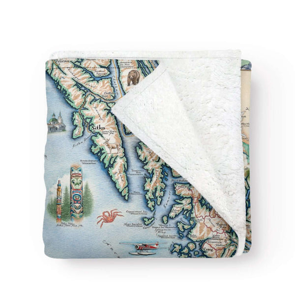 Folded Alaska's Inside Passage map fleece blanket throw in earth-tone colors. The map features totem poles of native peoples, bears, whales, fish, mountain goats & sheep. Flora is an illustration of forget-me-nots. Including the towns of Sitka, Juneau, Ketchikan, and others.