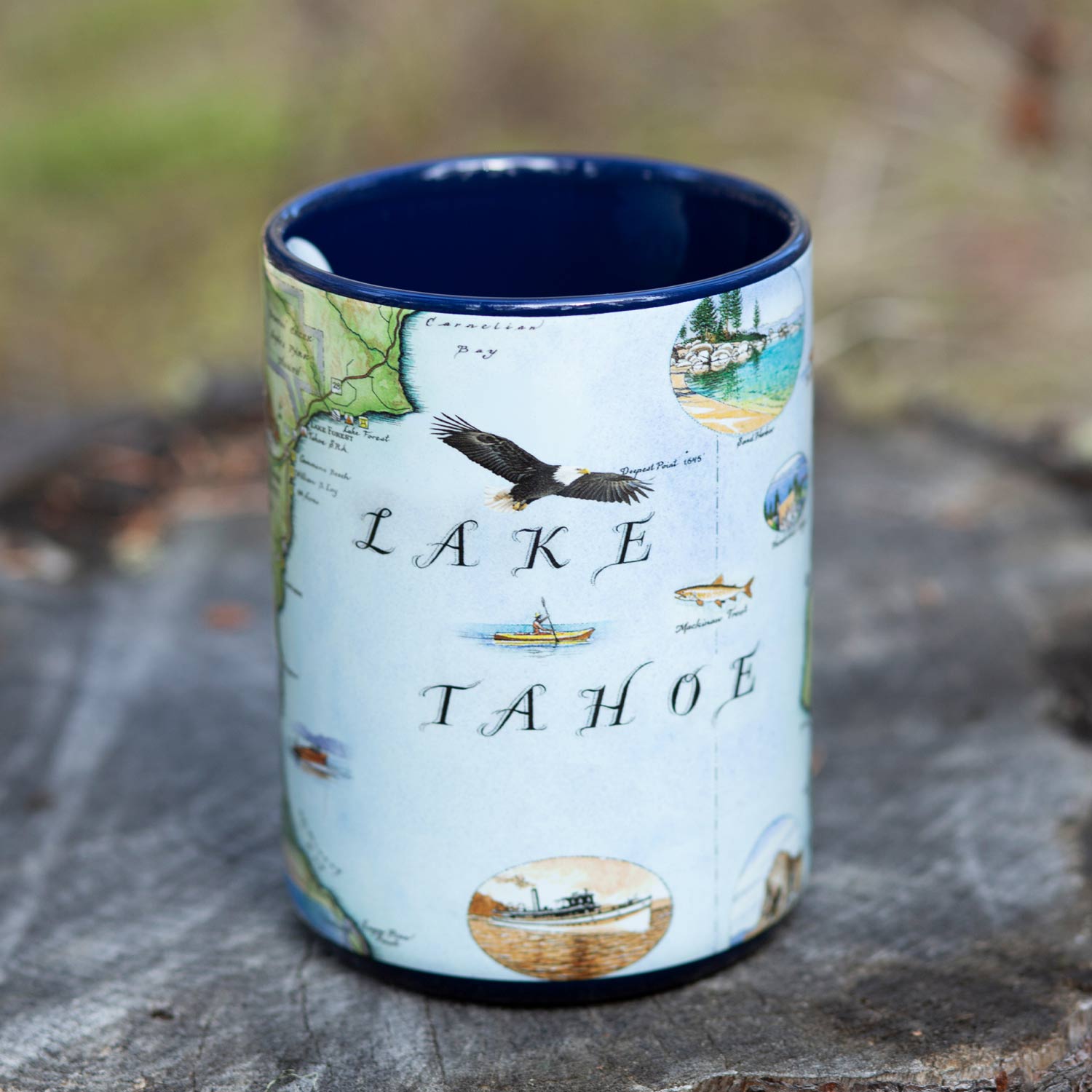 Blue 16 oz Lake Tahoe map ceramic coffee mug sitting on a tree stump. The cup features bald eagle, boats, bear, fish, Emerald Bay State Park, Spooner Lake, Cove Rock, and Thunderbird Lodge.