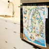 Michigan State Map Kitchen Dishwashing Towel hanging on the kitchen stove handle. The Map features the Great Lakes, Detroit, Ann Arbor, Grand Rapids, and Lansing in earth tones of blue and greens. The tea towel also features  Nature, animals, ducks, deer, fish, moose lighthouses, wolverines, and the Mackinac Bridge.