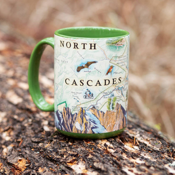Green 16 oz North Cascades National Park map ceramic coffee mug. The cup features Mountain bikers, flowers, Ross Lake Recreation Area, and Majestic Mountains.