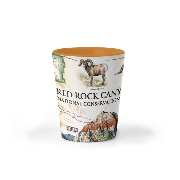 Red Rock Canyon National Park Map Ceramic shot glass by Xplorer Maps. The map features illustrations of places such as Mt. Willson, Rainbow Mountain, Bridge Mountain, and Spring Mountain Ranch. Flora and fauna include a red tail hawk, desert Bighorn, Globemallow, Joshua tree, and Prickly pear cactus.